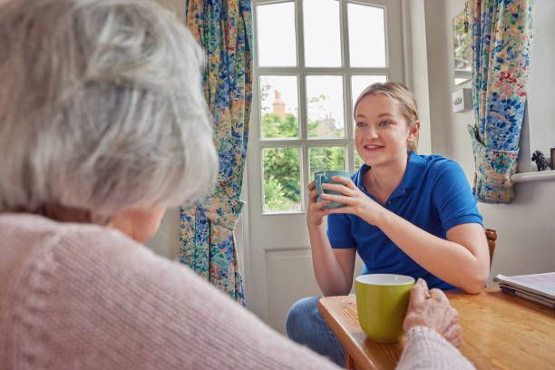 Woman in blue shirt chats with a person seated across a table, both holding mugs in a room with a window.