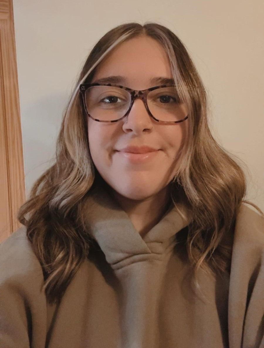 Woman with glasses, wearing a tan hoodie, facing the camera.  Light brown hair with highlighted sections. Neutral expression.