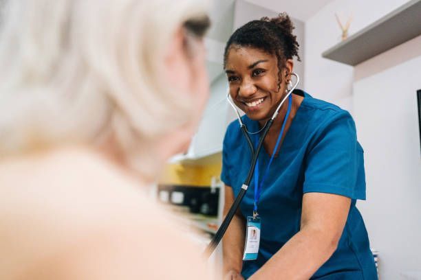 Nurse in blue scrubs listening to a patient's chest with a stethoscope. She is smiling.
