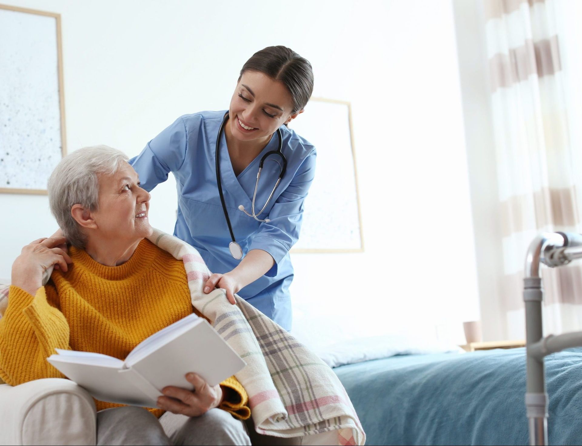 Nurse putting a blanket on an elderly woman sitting and reading a book. Indoor setting.
