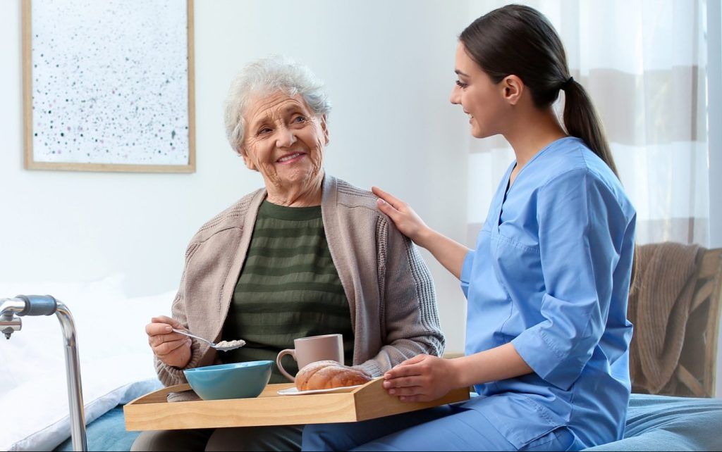 A caregiver serves breakfast to an elderly person in bed, smiling and holding a tray.