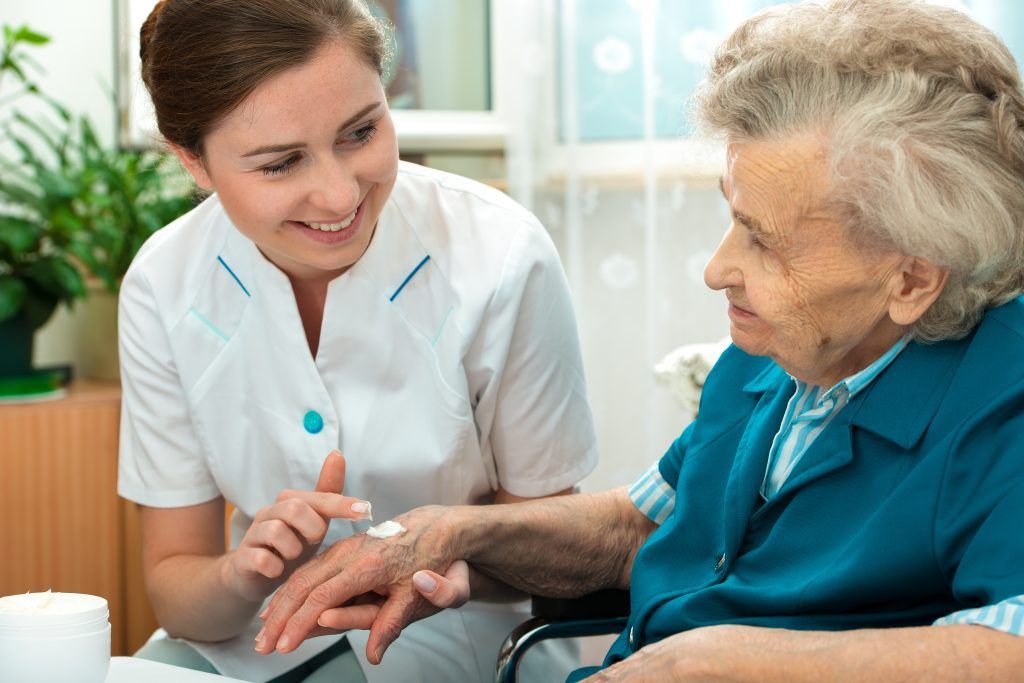 Caregiver applying cream to an elderly person's hand. Indoor setting, plants in background.