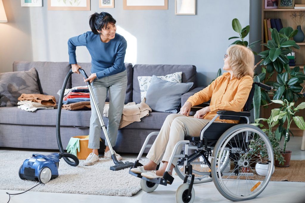 Woman vacuums rug, another sits in wheelchair, talking in living room.