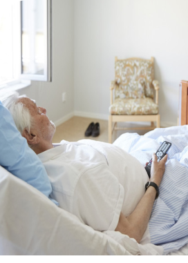 Person in bed holding a remote, looking at the TV. The room has a window and a chair.