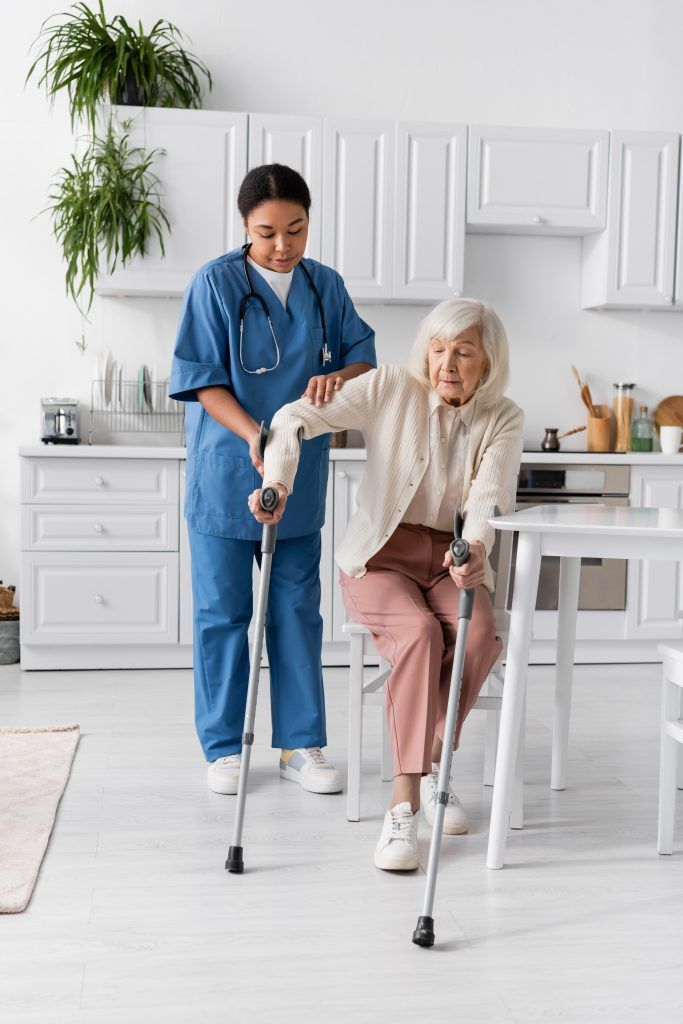 Nurse assisting a person using crutches in a kitchen, supporting them while they walk.