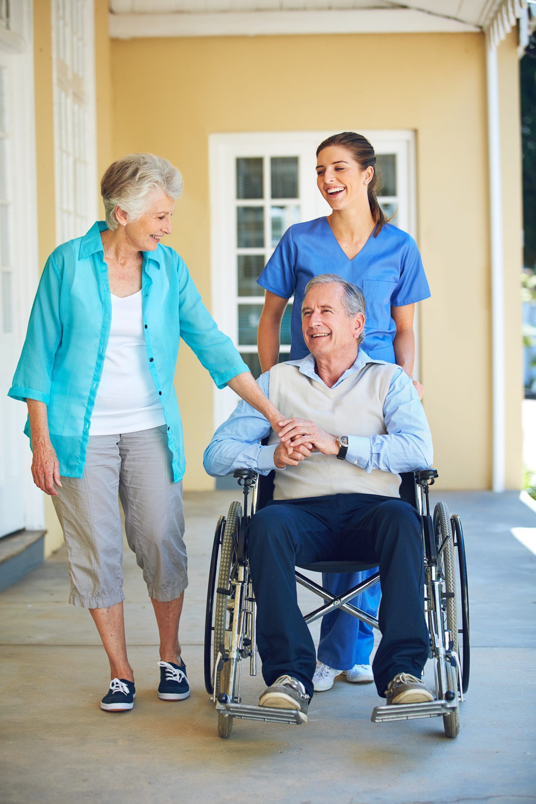 Elderly man in wheelchair, woman walking beside, nurse pushing. All smiling on porch.
