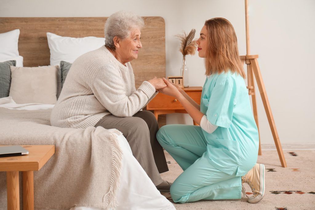 Caregiver kneeling beside senior on a bed, holding her hands and talking. Light, neutral setting.