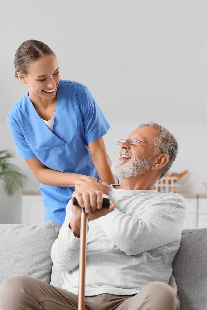 Caregiver assists a person with a cane sitting on a couch. Both are smiling.