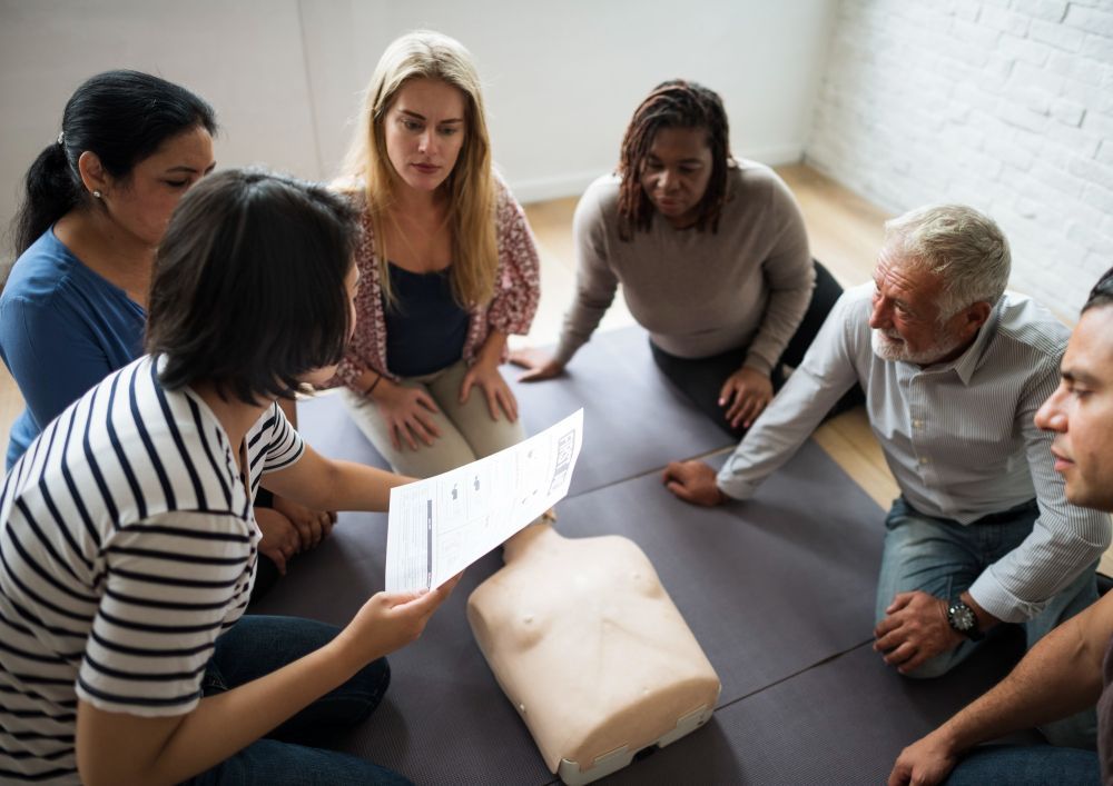 People in a CPR training, gathered around a mannequin and a trainer reading instructions.