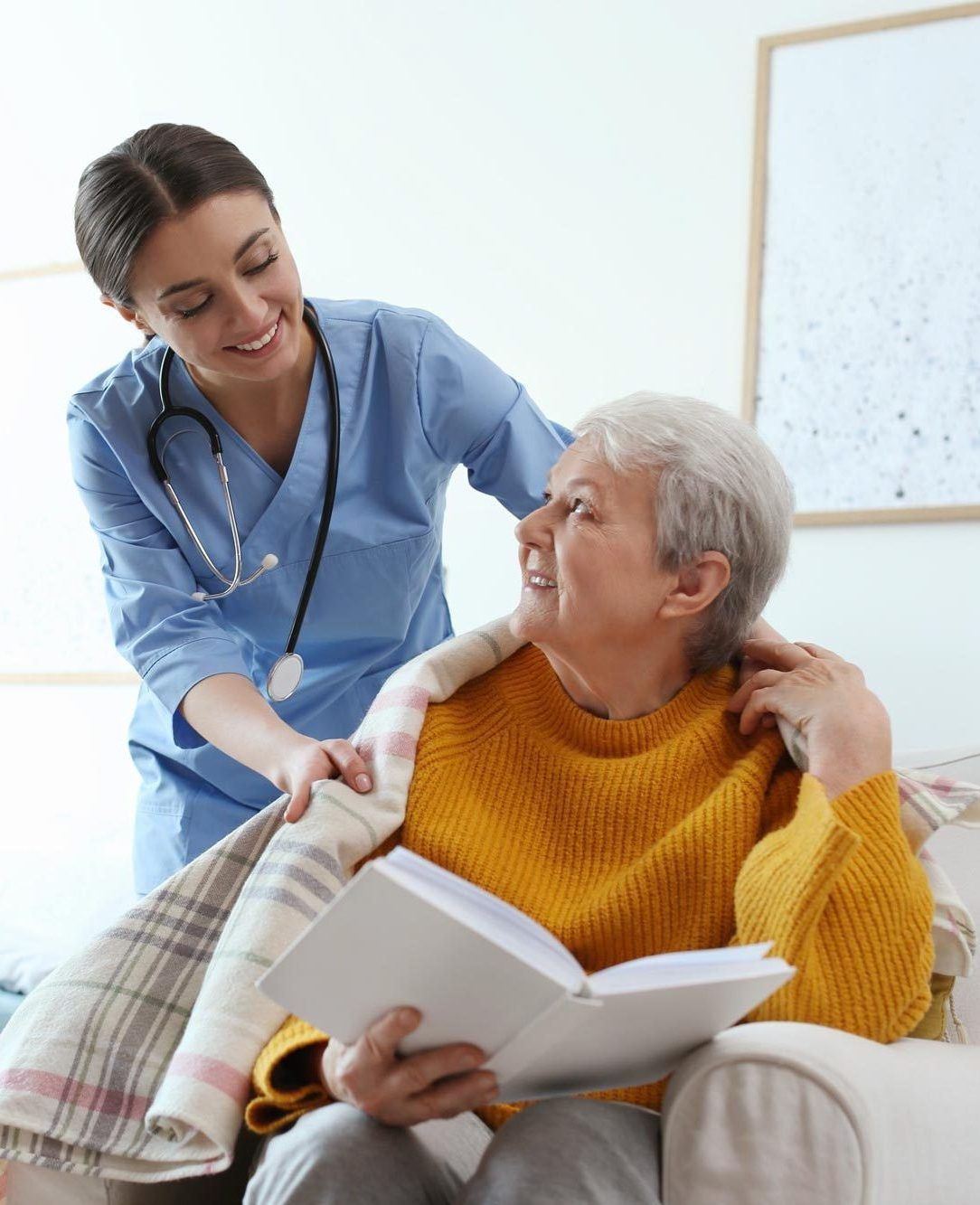 Nurse comforts senior reading, indoors. Nurse in blue scrubs, senior in yellow sweater, both smiling.
