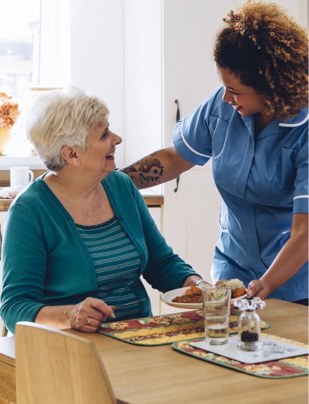 Woman eating at a table, caregiver serves her a meal.
