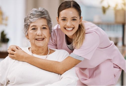 Woman in pink scrubs embraces an older woman, both smiling, indoor setting.