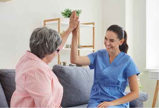Woman in blue scrubs high-fives an older person on a couch. Both are smiling inside a living room.