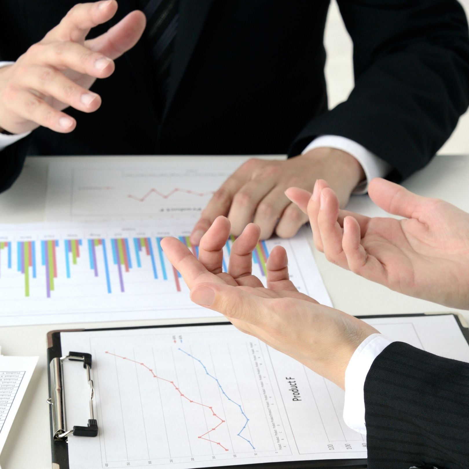 Hands discussing business charts and graphs on a clipboard at a meeting table.