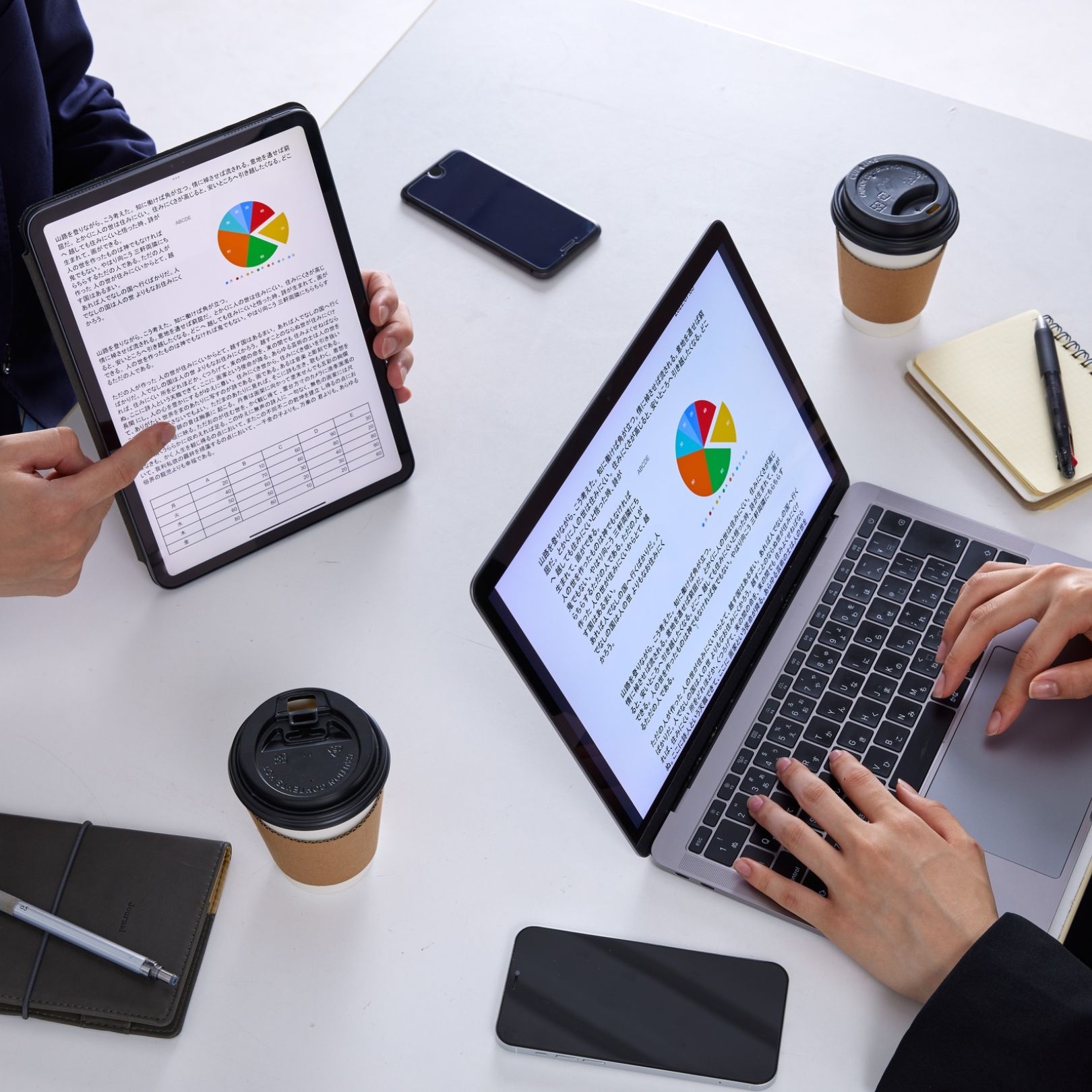 Two people reviewing charts on a tablet and laptop at a desk with coffee cups and phones
