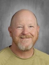 A bald man with a beard is wearing a yellow shirt and smiling.