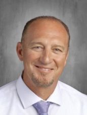 A man in a white shirt and purple tie is smiling for the camera.