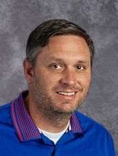 A man in a blue shirt and purple and red striped shirt is smiling for the camera.