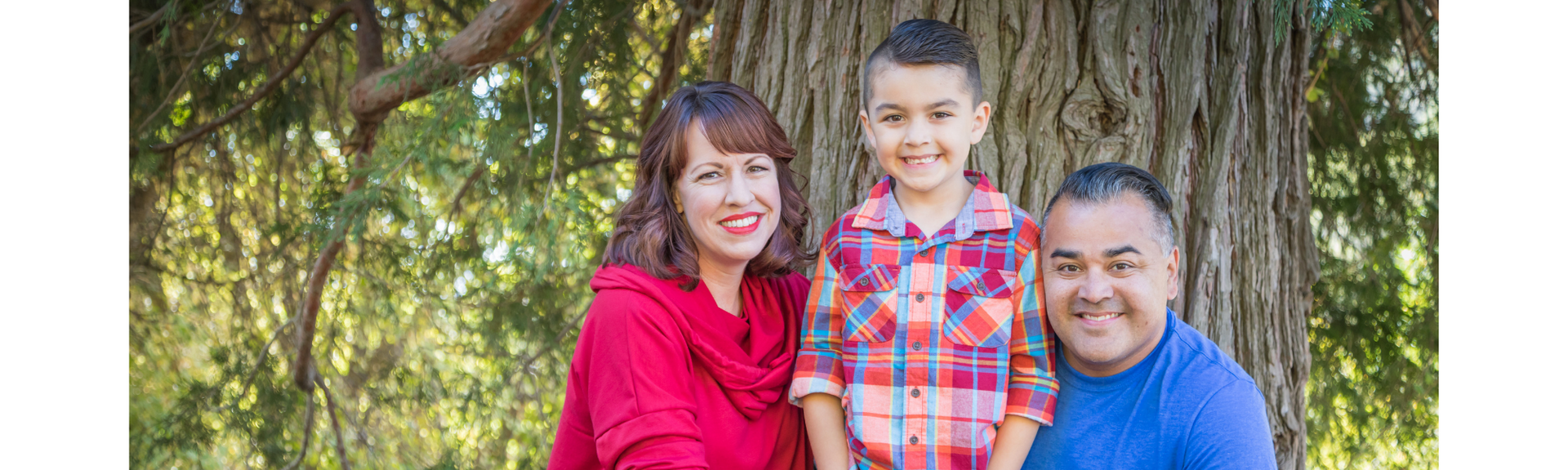 Three smiling people standing outdoors in front of a tree, with one child in the middle.
