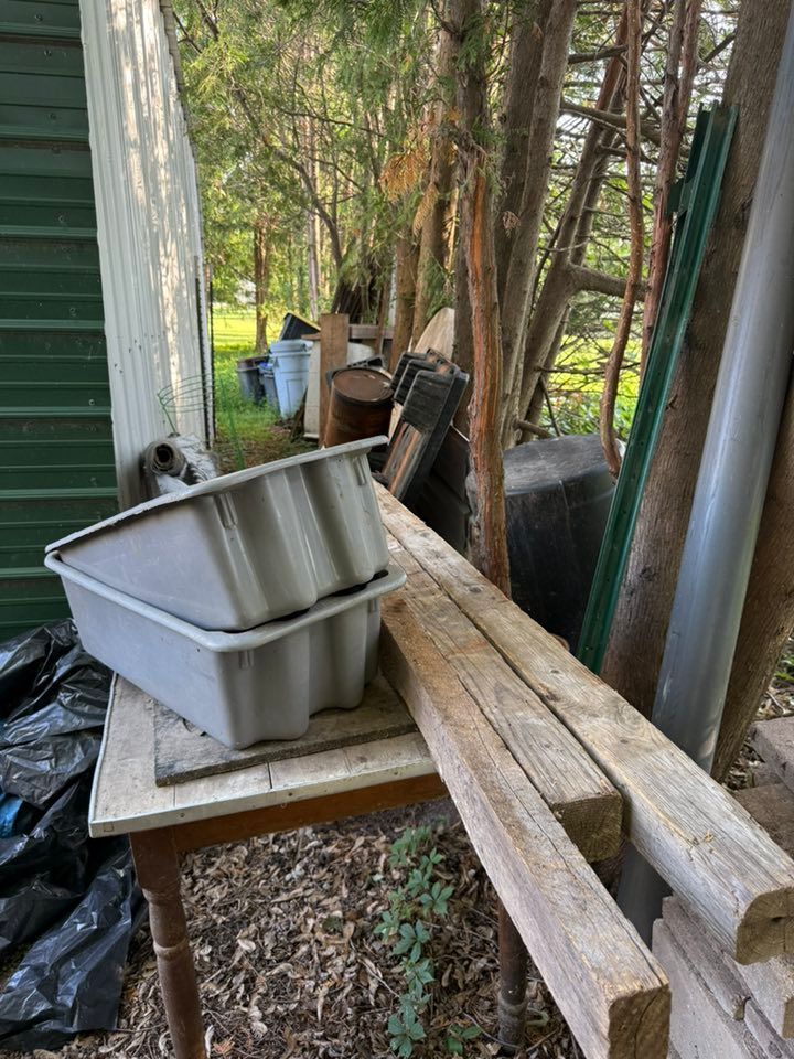 Two light gray plastic bins stacked on a wooden table outdoors, with lumber resting nearby and trees in the background.