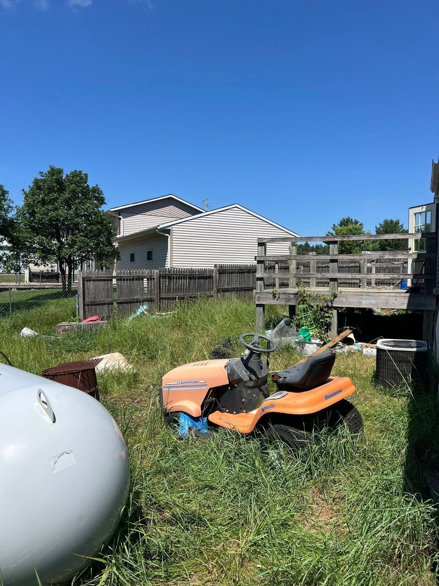 An orange riding lawnmower sits in an overgrown grassy yard near a propane tank and a wooden deck under a clear blue sky.