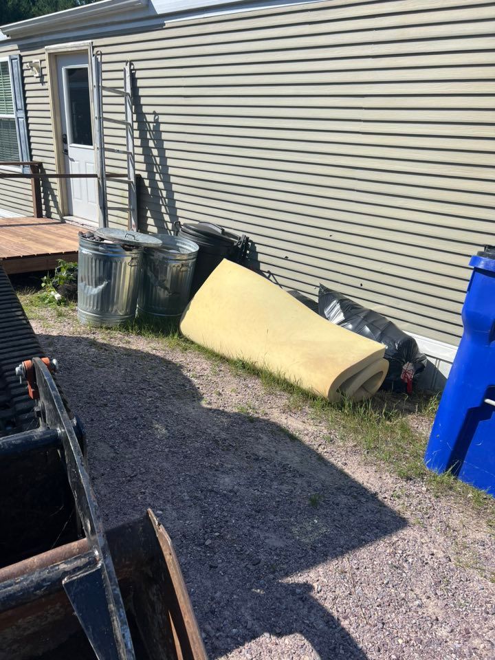 A rolled-up yellow foam mattress pad and two metal trash cans sit on a gravel driveway beside a tan-sided mobile home.