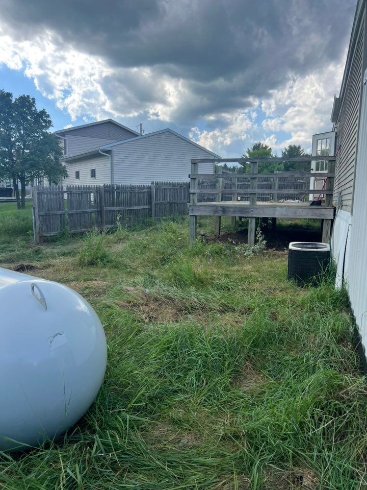 A grassy backyard with a wooden deck, a propane tank in the foreground, an AC unit, and a wooden fence under a cloudy sky.