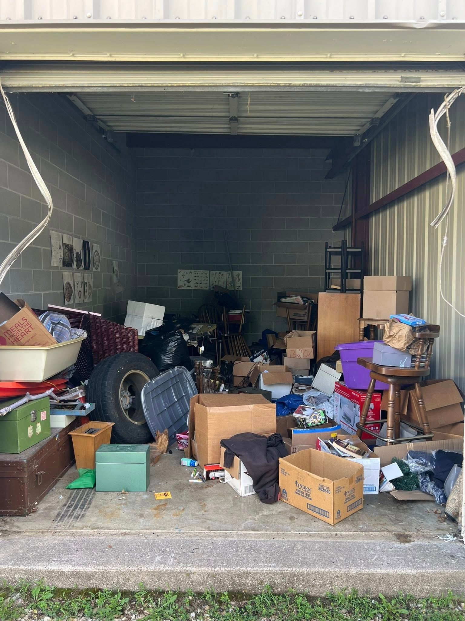 A cluttered, open storage unit filled with cardboard boxes, a car tire, furniture, and miscellaneous household items.