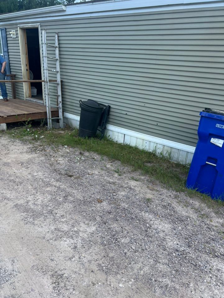 A light green mobile home exterior with a wooden porch, an open door, a leaning ladder, and two waste bins on gravel.