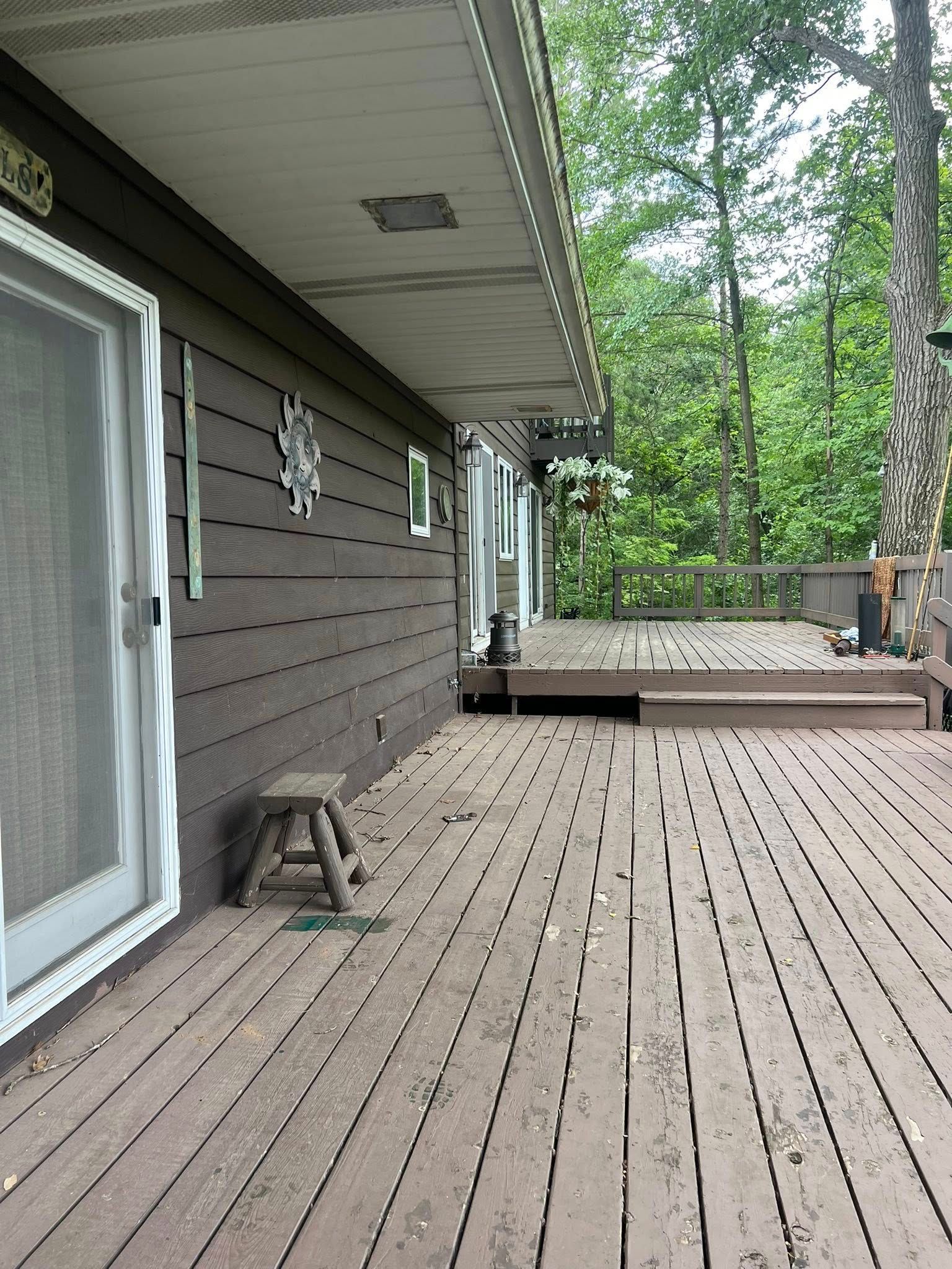 A wooden deck extends from the side of a house toward trees, with a small wooden stool sitting near a glass sliding door.