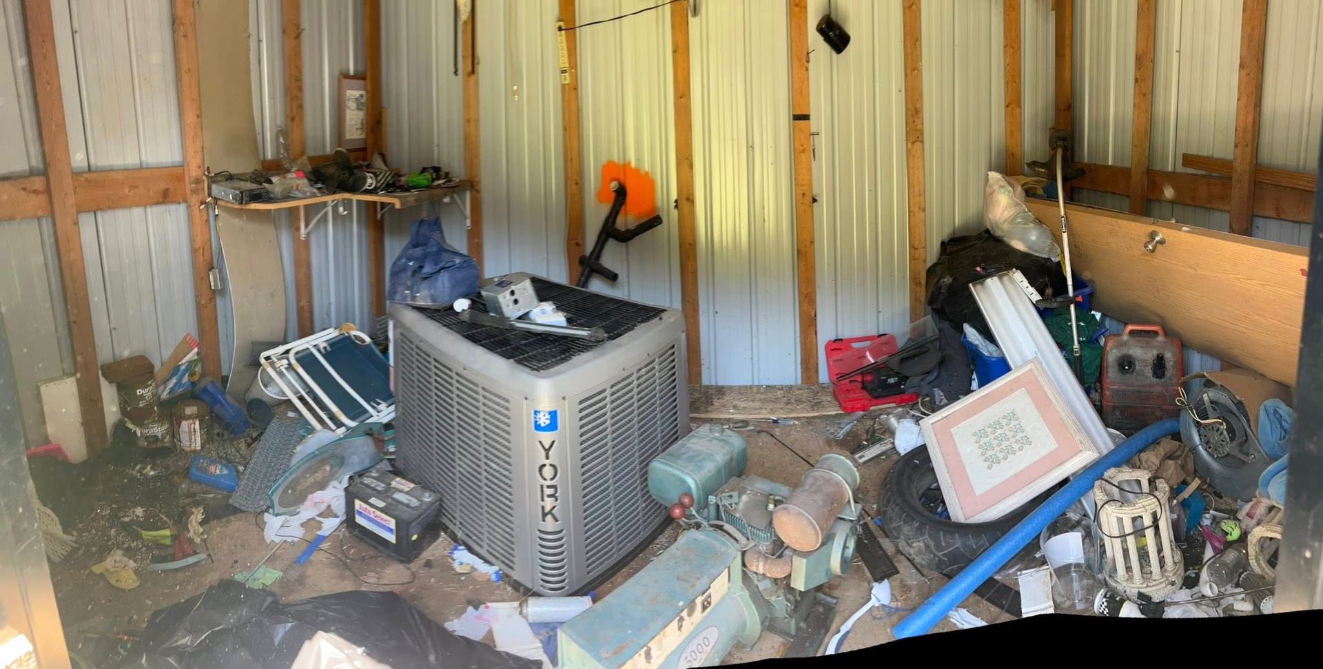 A cluttered shed interior featuring a large metal AC unit, tools, and various scattered household items.