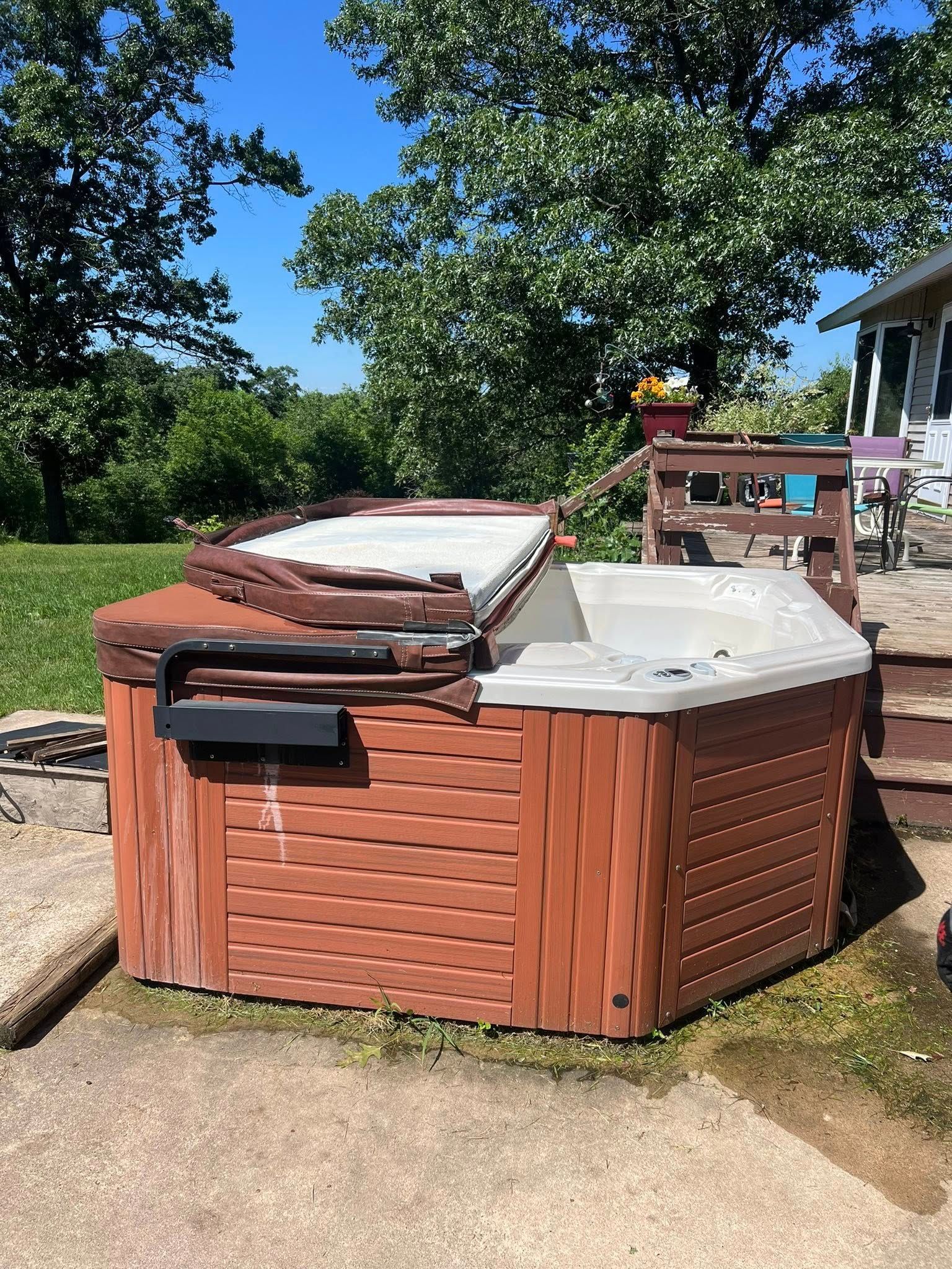 A brown, rectangular hot tub with its cover partially open, situated outdoors on a concrete patio near a grassy area.