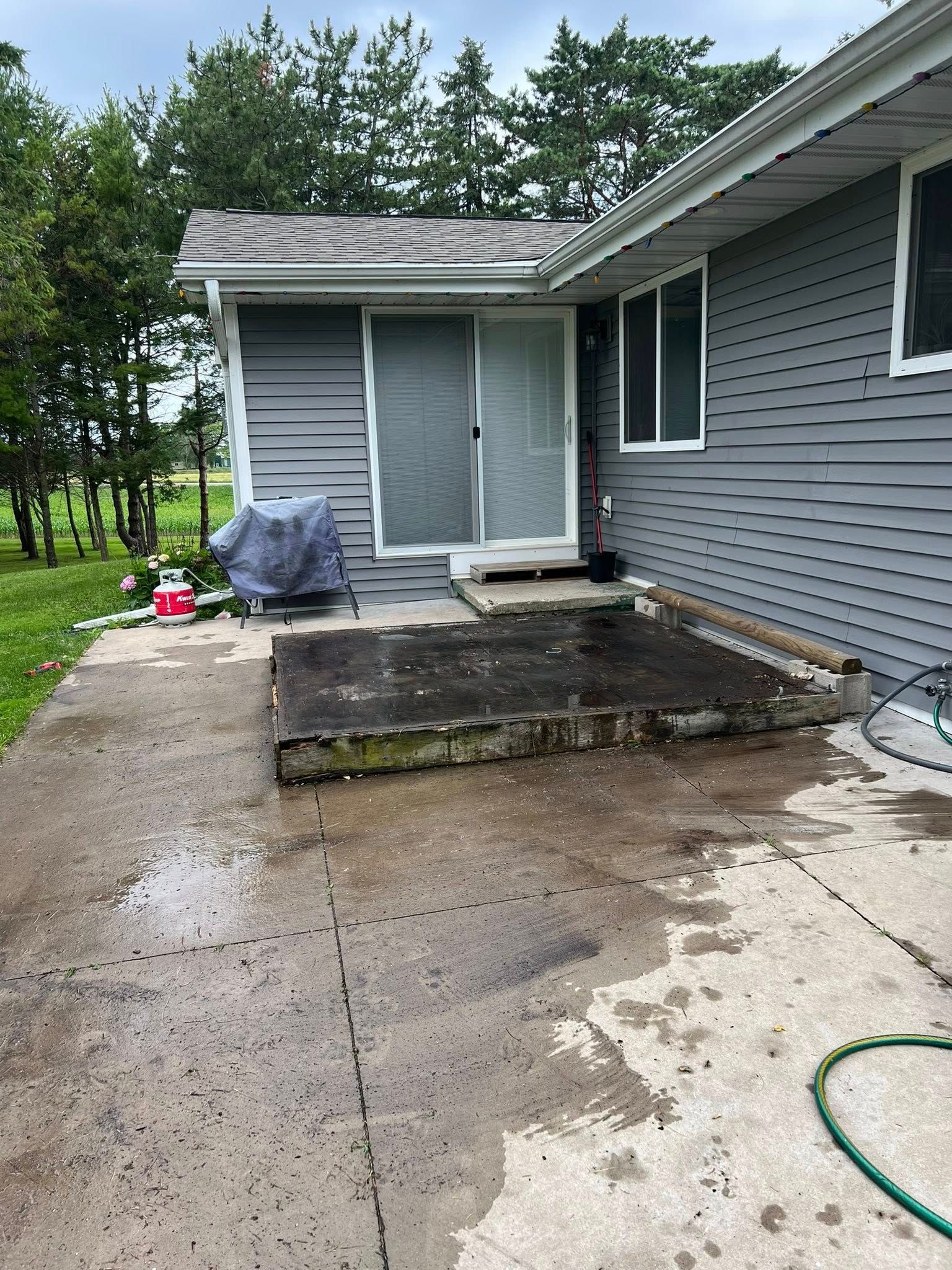 Gray house exterior with a sliding glass door, a raised dark concrete platform, and a grill cover on a wet patio.