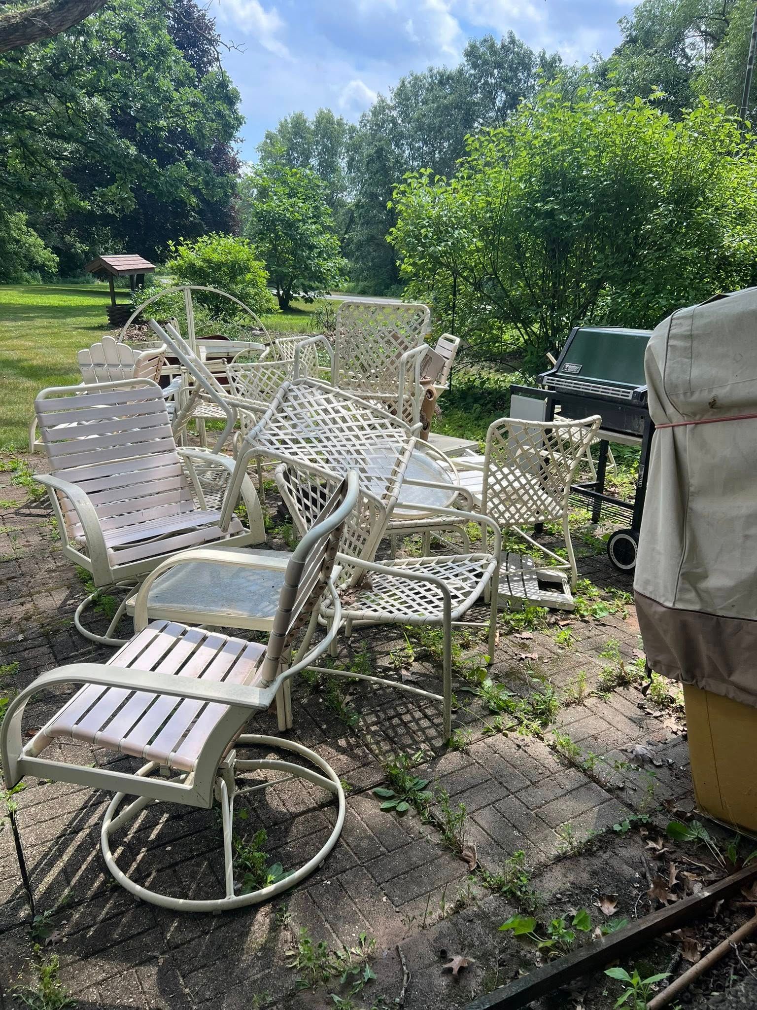 A group of weathered white metal outdoor chairs and a covered grill sit on a patio surrounded by trees and greenery.