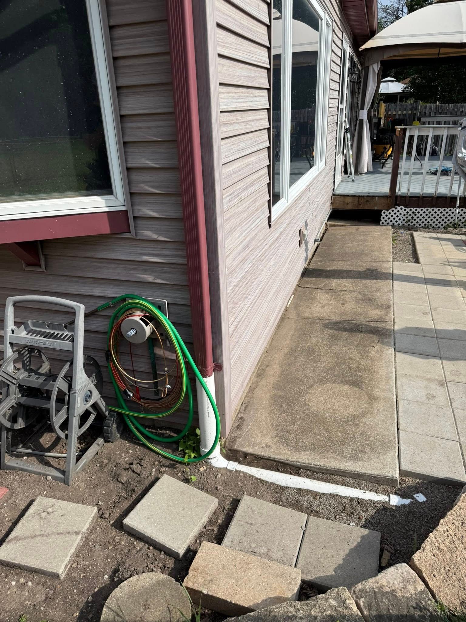 A side view of a house exterior with beige siding, a green garden hose, a concrete walkway, and stone pavers on the ground.