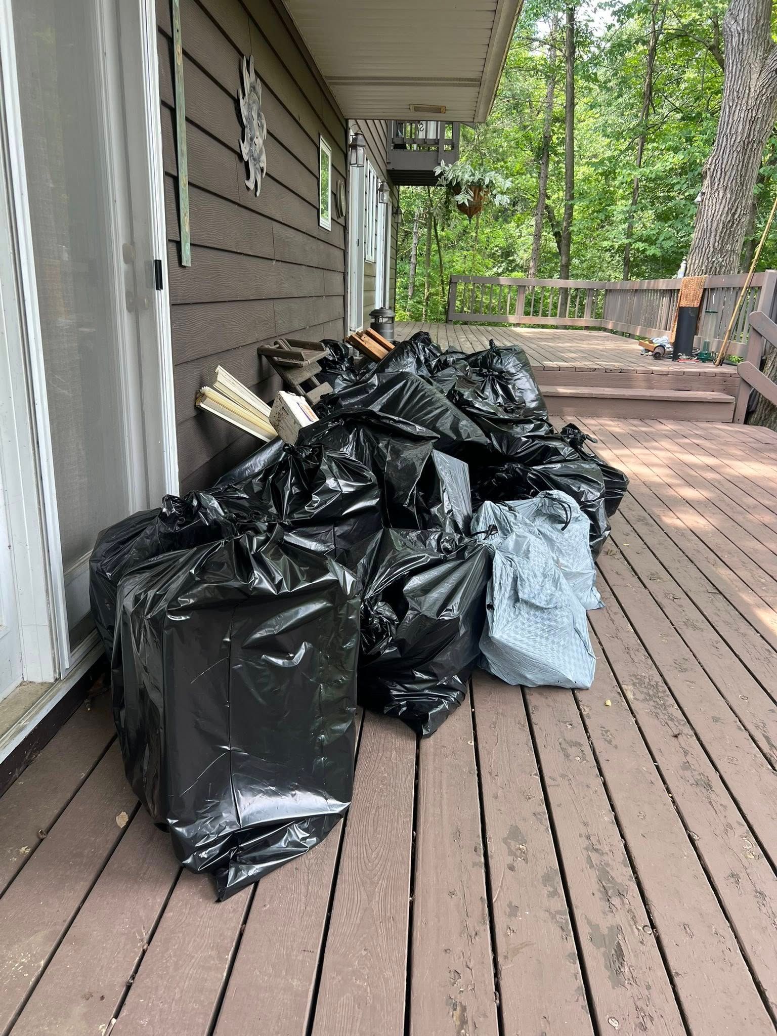 A large pile of black trash bags and some loose lumber on a wooden deck next to a house exterior.