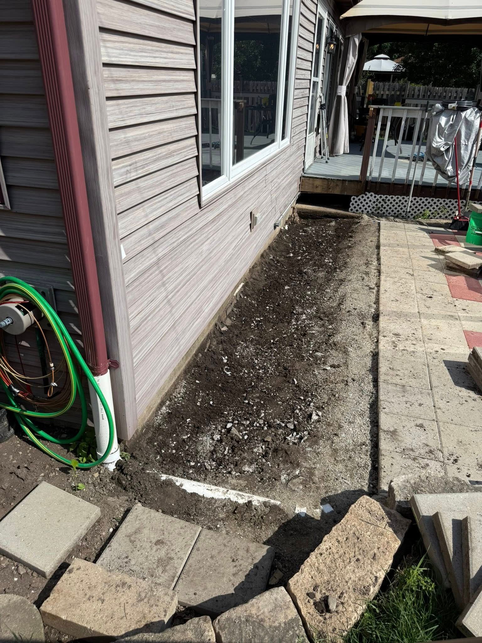 A freshly tilled dirt garden bed runs along the side of a house next to a concrete patio and stone border.