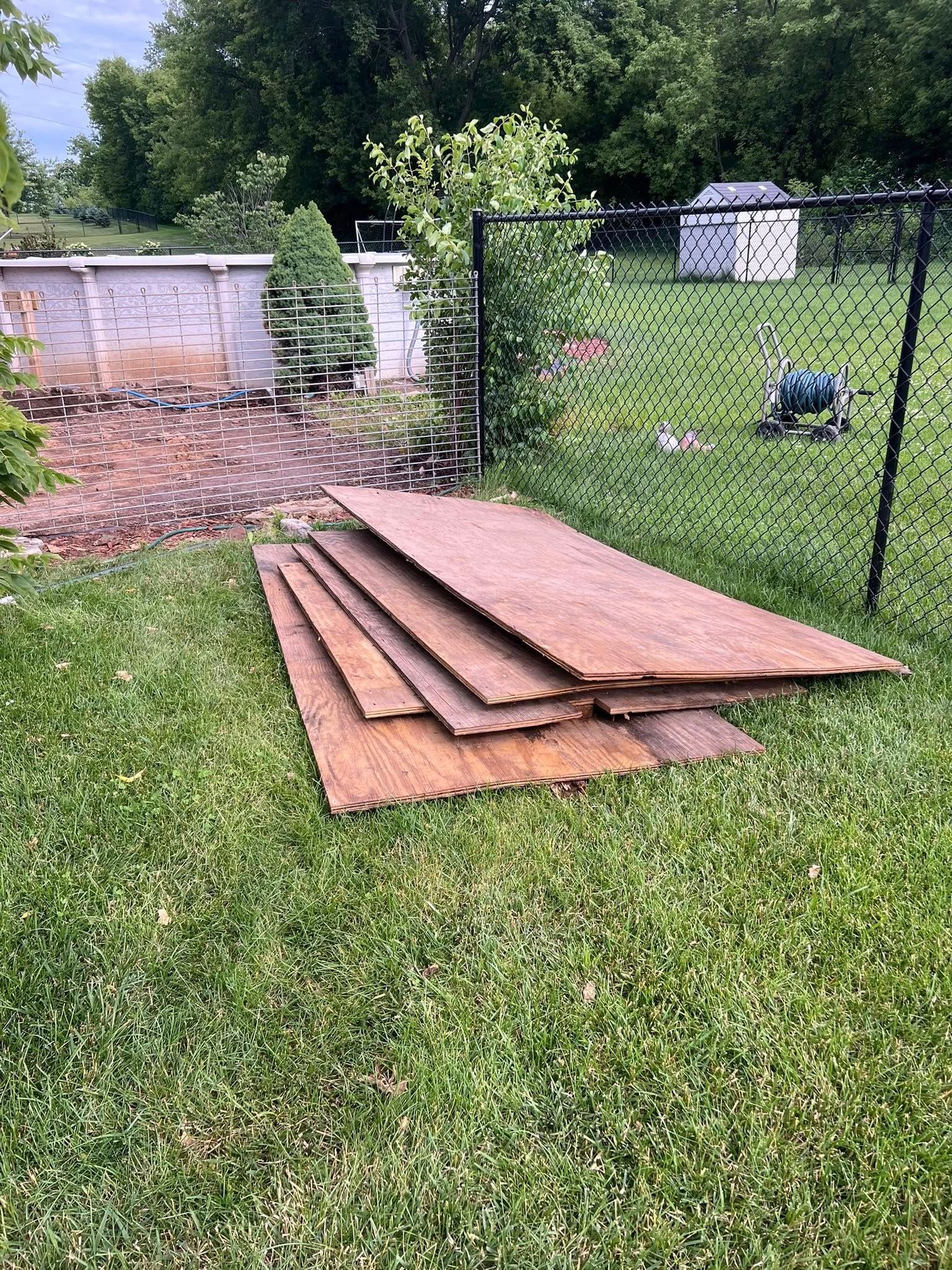 A stack of several rectangular wooden boards lying on the grass near a chain-link fence and an above-ground pool.