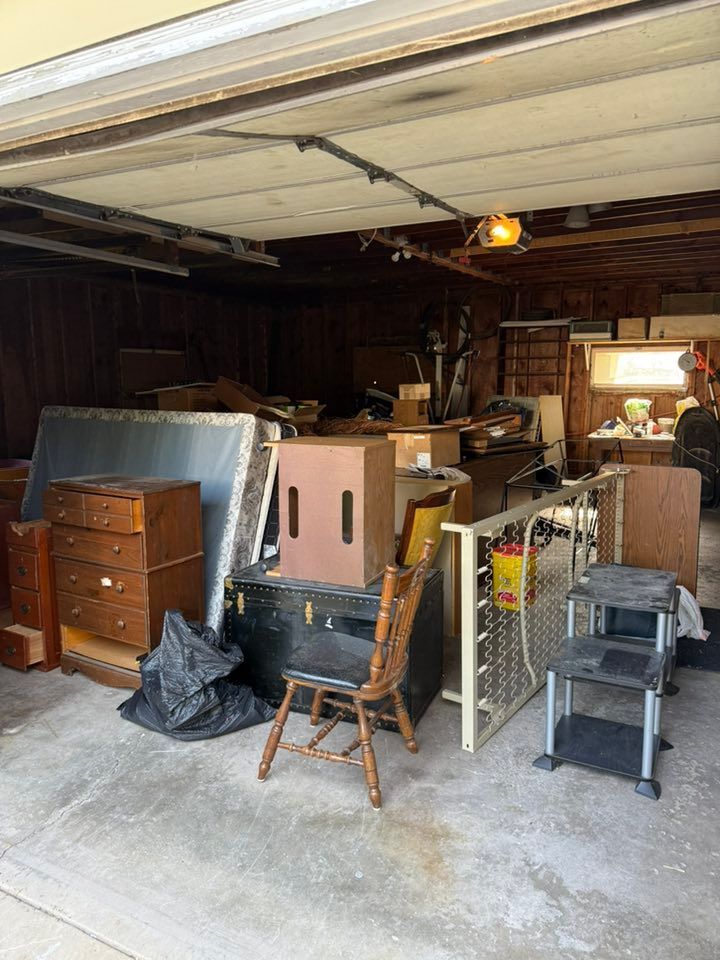 A cluttered residential garage filled with furniture, including a dresser, mattress, chairs, and miscellaneous items.