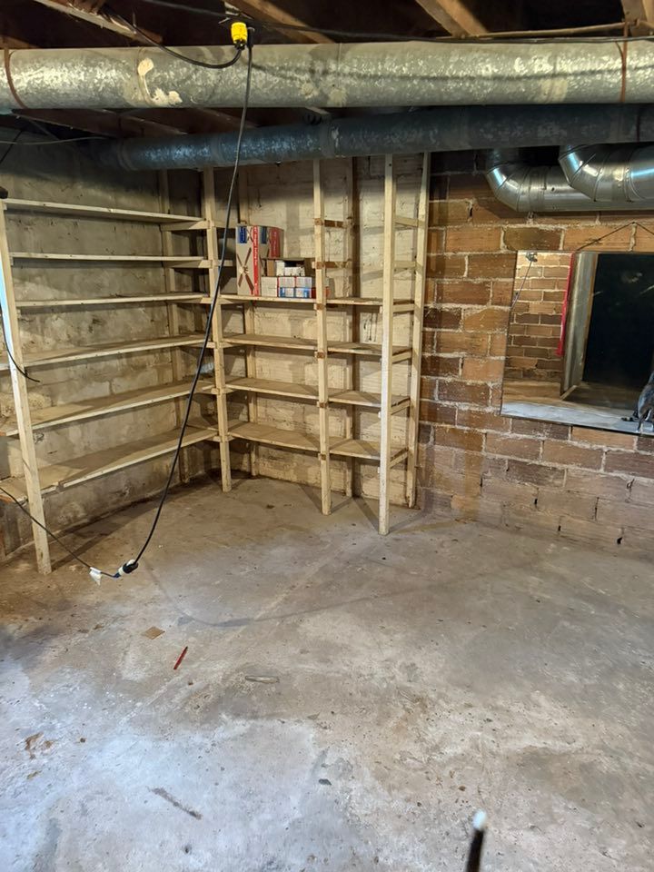Wooden shelving units stand in the corner of a dimly lit, unfinished basement with exposed brick walls and ductwork.