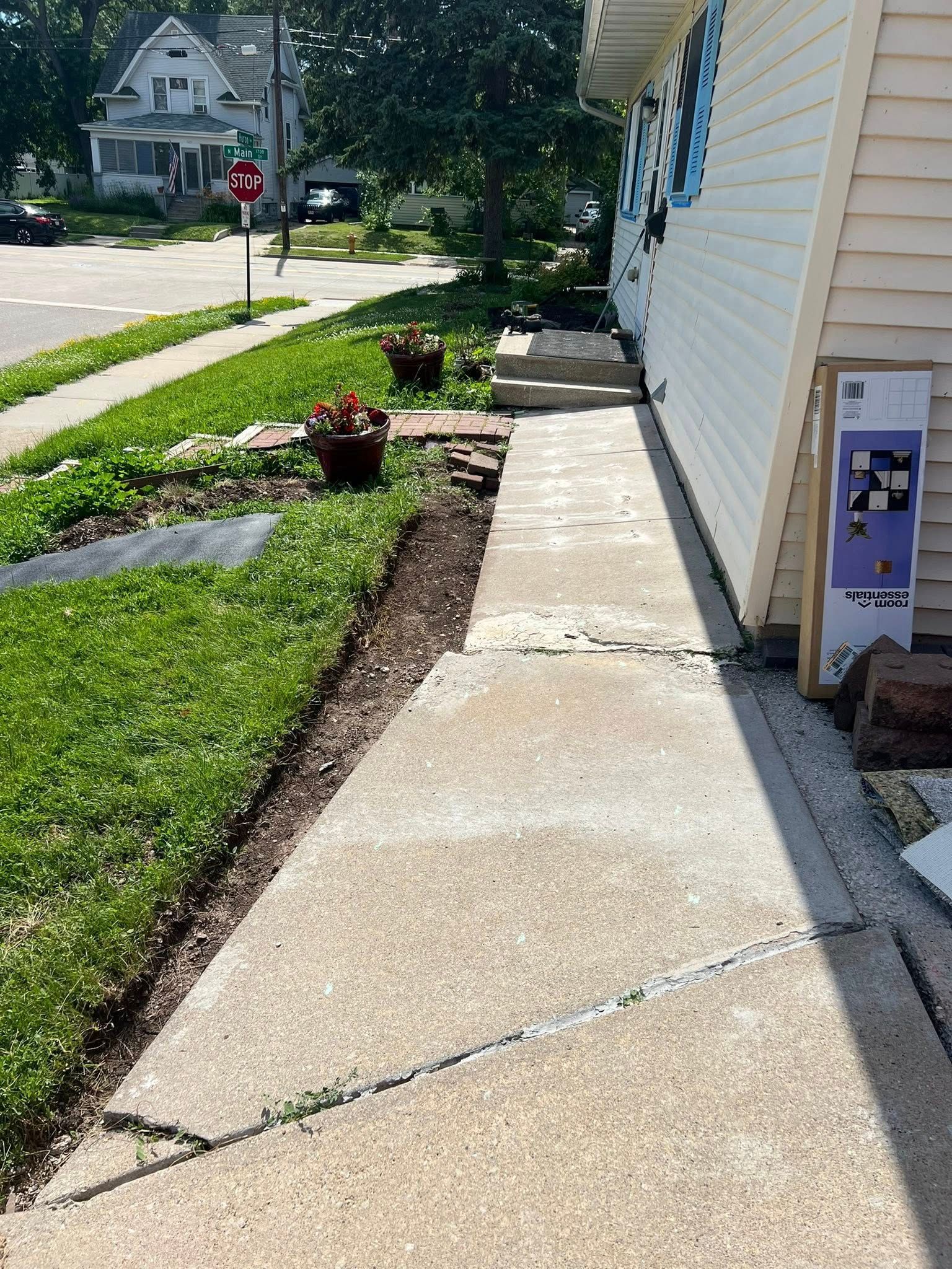 A sidewalk leads to house steps, with a cracked concrete path and a decorative board leaning against the building.