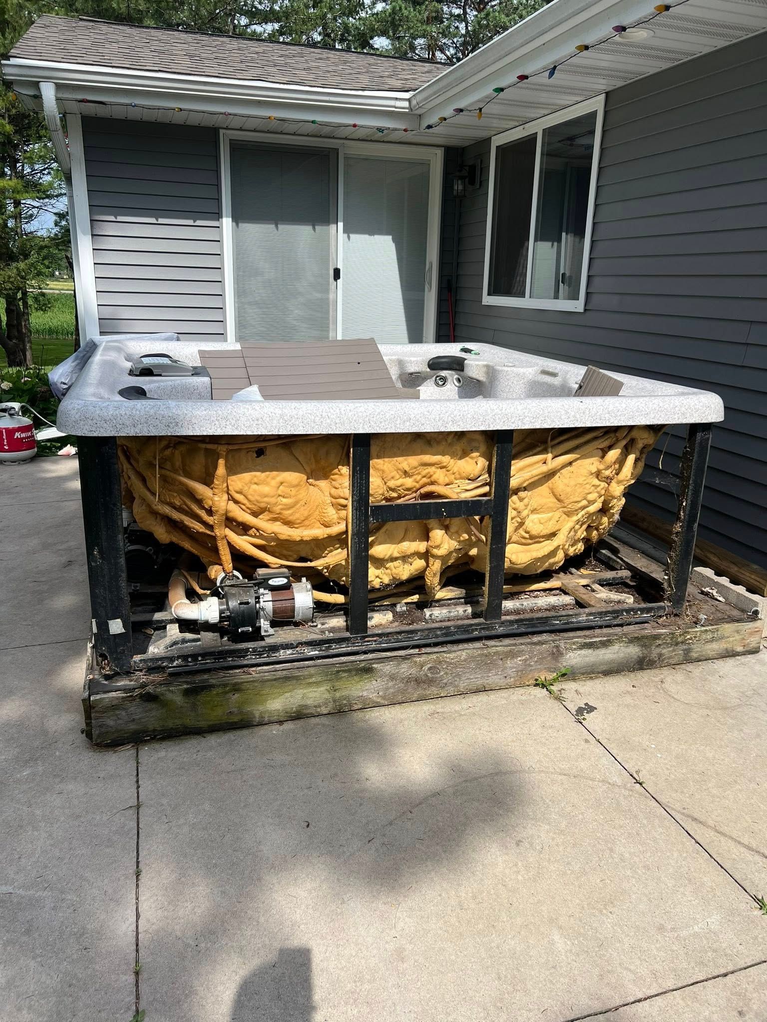 A hot tub with its side panels removed, revealing yellow spray foam insulation and internal plumbing on a concrete patio.