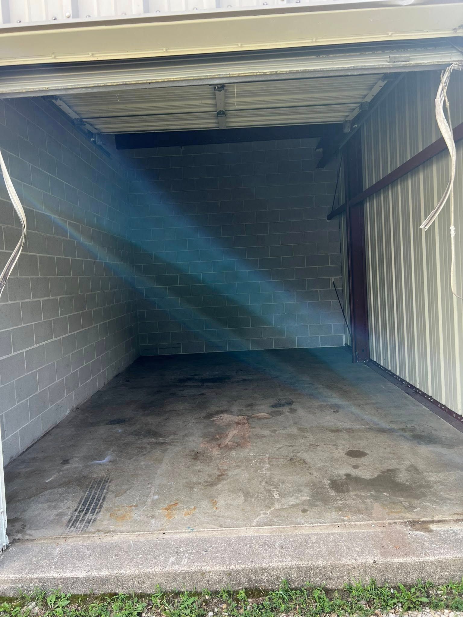 A view into an empty storage unit with a gray concrete floor, light-colored brick walls, and a metal roll-up door.