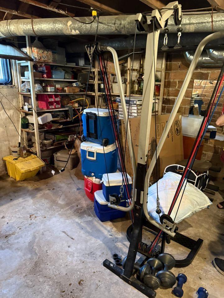 A gym machine with resistance bands stands in a cluttered basement with storage shelves and a stack of colorful coolers.
