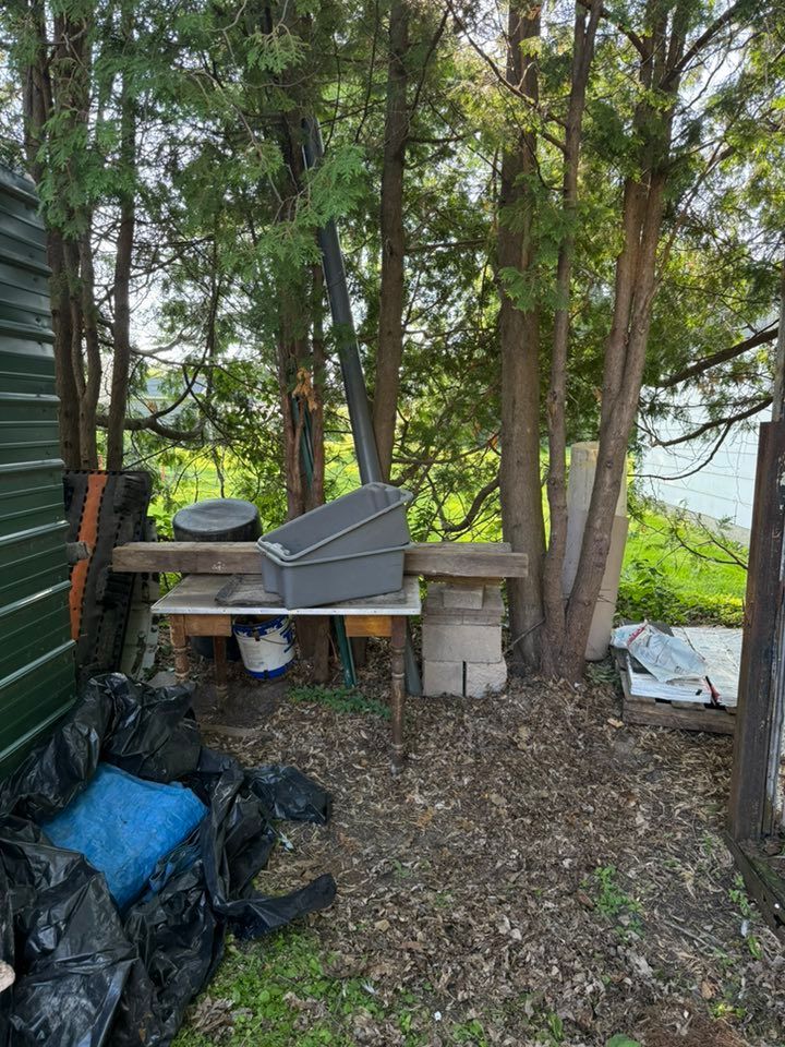 A makeshift table holds plastic bins and a wooden beam near a green shed and trees in a shaded yard.