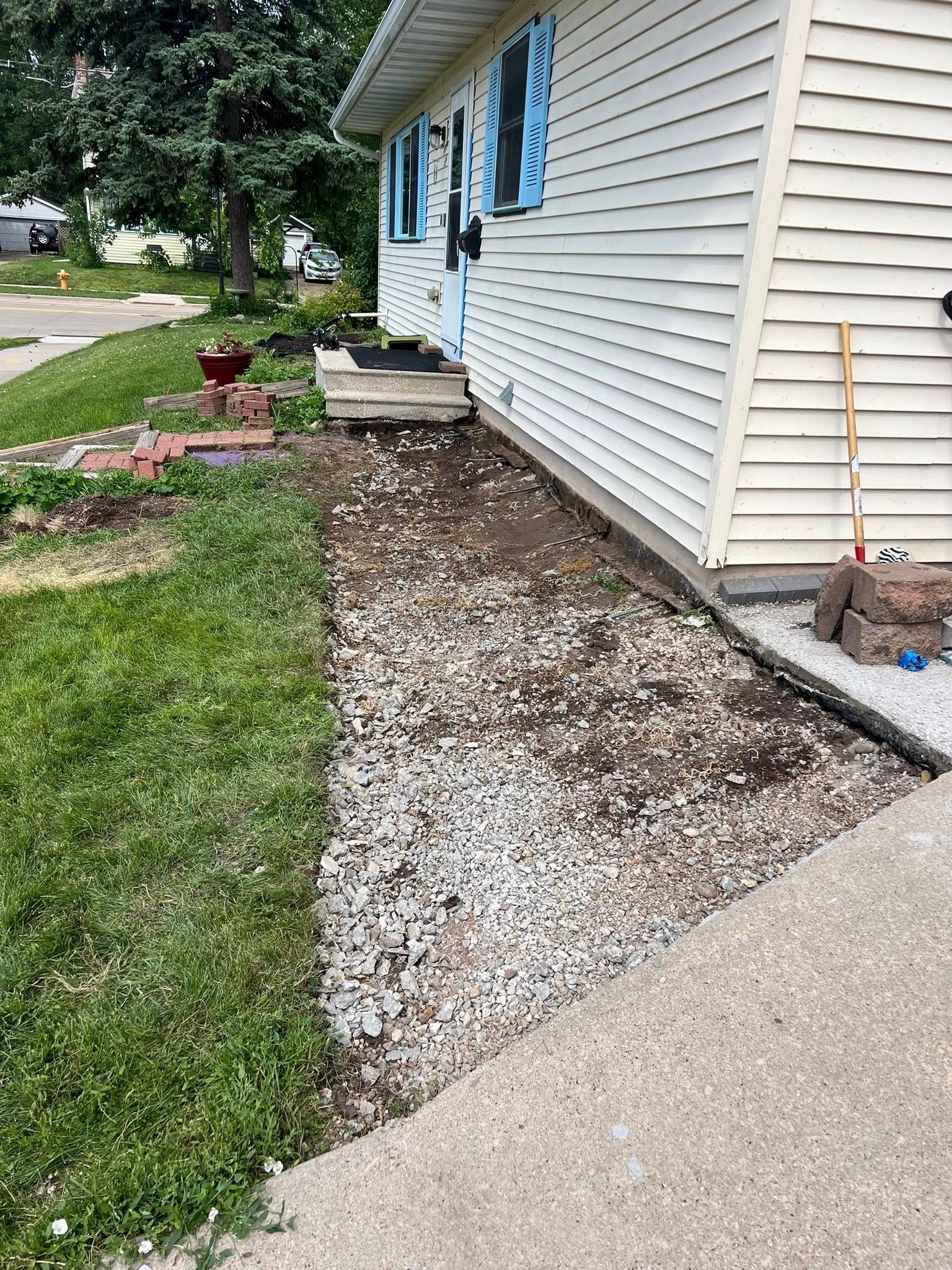 A side yard with a gravel pathway along a white-sided house, leading toward a small wooden porch and stairs.