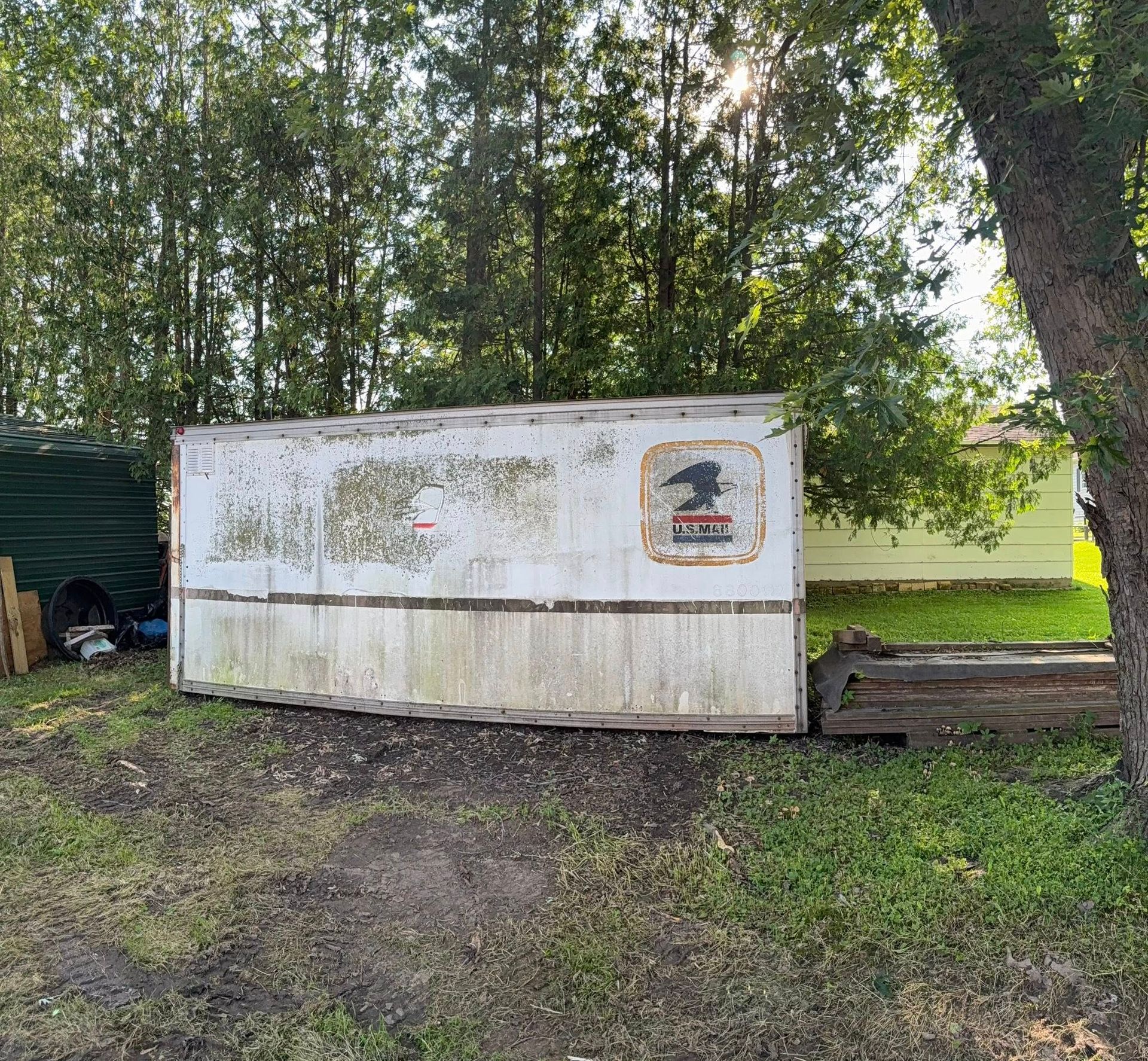 An old, weathered white shipping container with a faded U.S. Postal Service logo, sitting in a grassy, tree-lined yard.
