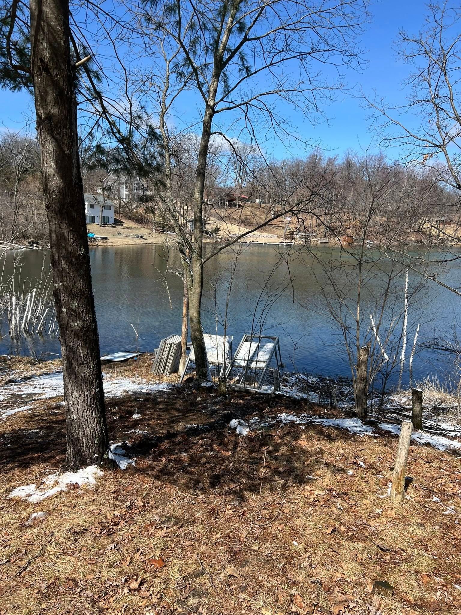 Two white folding chairs sitting by a calm lake on a sunny day with patches of snow on the ground.