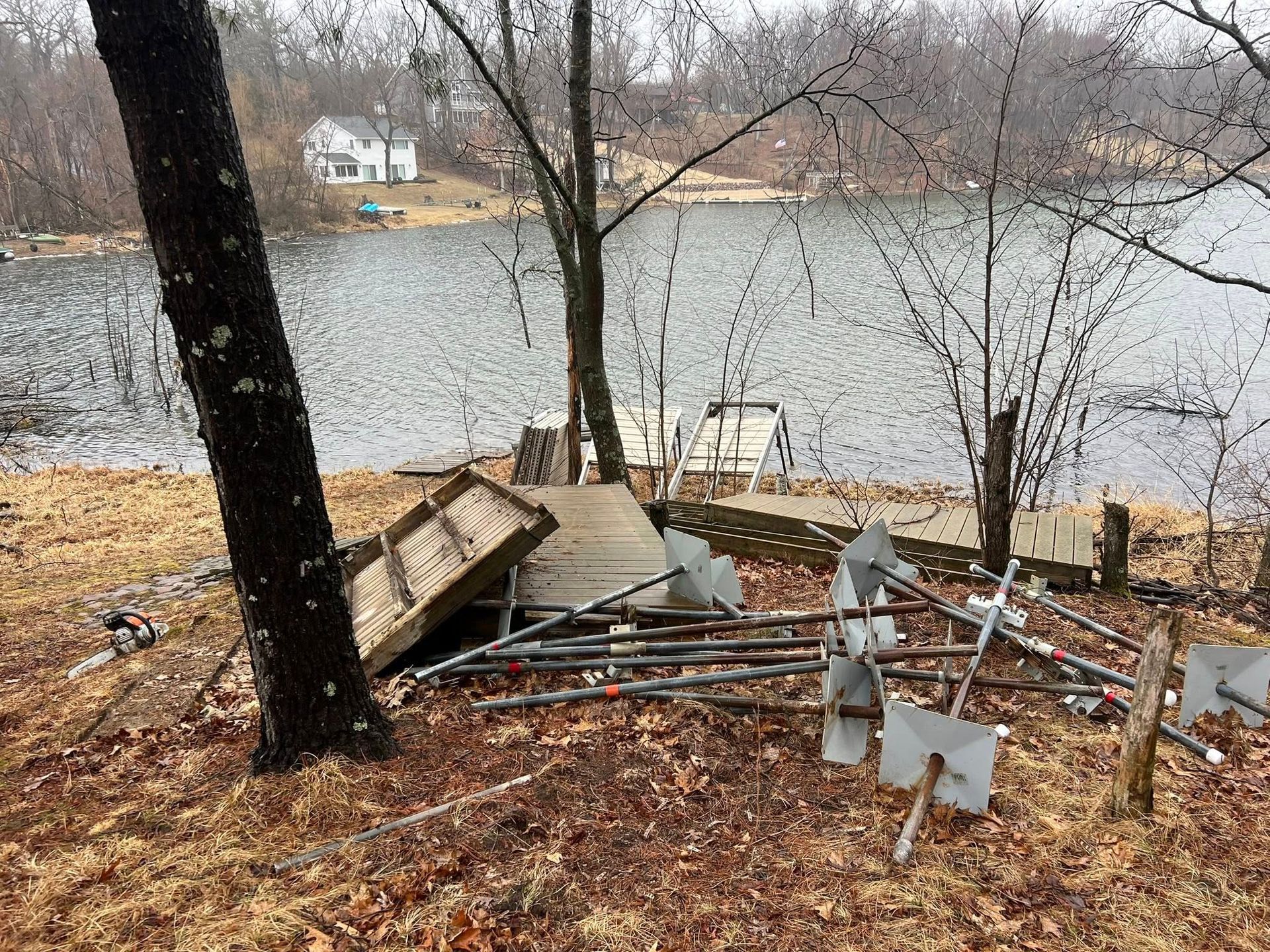A lakeside yard featuring a dock section and metal poles lying on the ground near a large tree, facing a body of water.