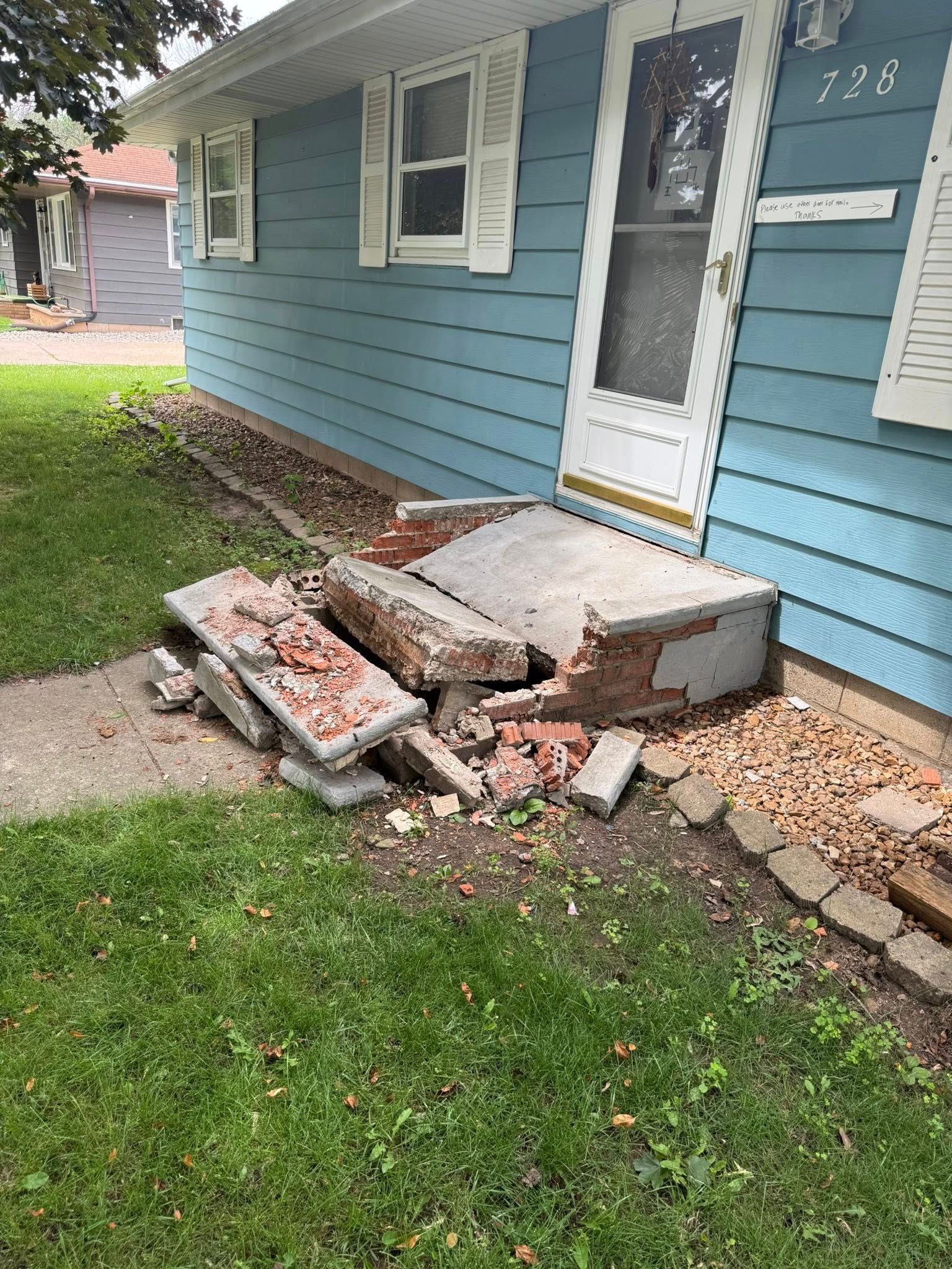 A blue house with a collapsed concrete front porch step and broken masonry on a grassy lawn.